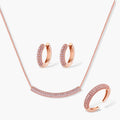 Rose gold jewelry set with pavé cubic zirconia stones including curved necklace, hoop earrings, and adjustable ring on cream background.