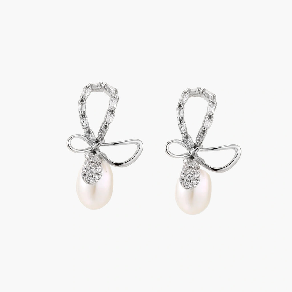 Silver bow-shaped stud earrings with baguette crystals and dangling white freshwater pearls on cream background
