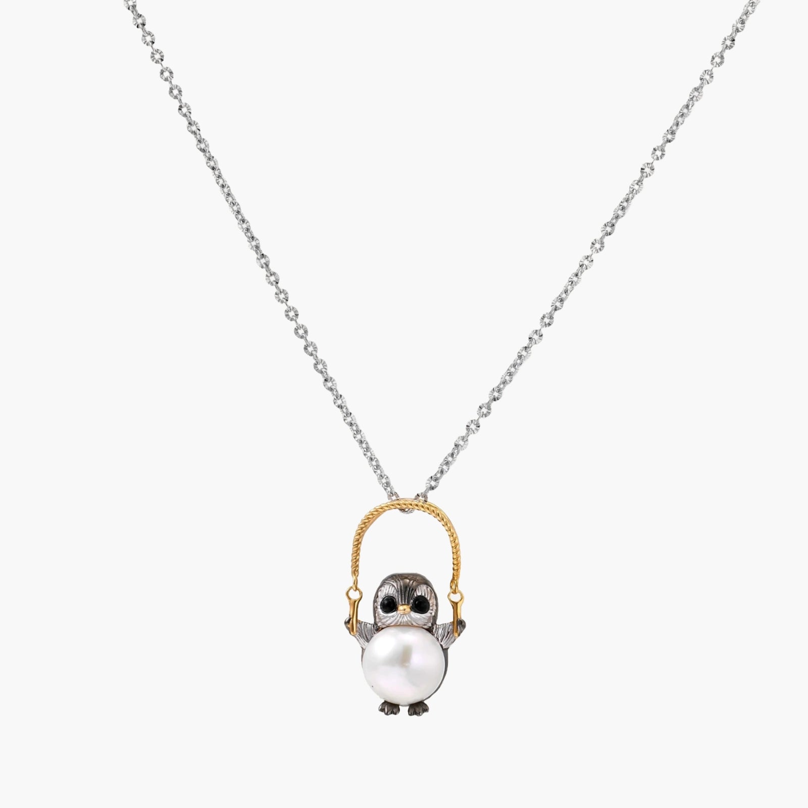 Whimsical owl pendant holding a white pearl on a gold swing, rhodium-plated silver chain on cream background.