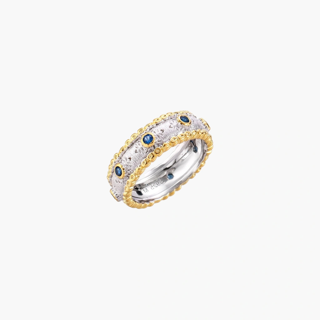 Two-tone ring with brushed silver center, gold beaded edges, and blue zirconia stones on white background.