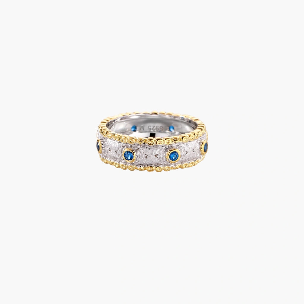 Two-tone ring with brushed silver center, gold beaded edges, and blue zirconia stones on white background.